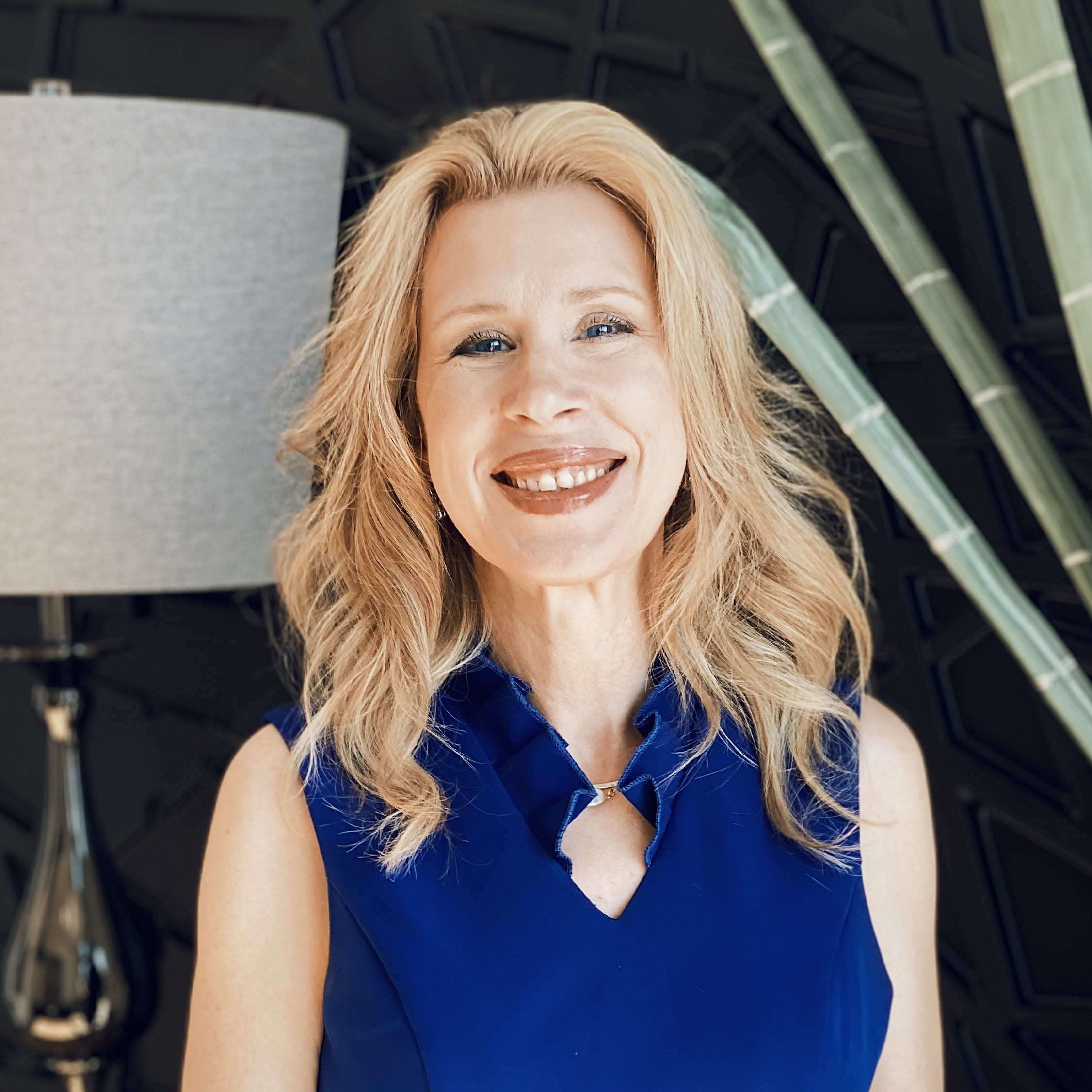A smiling woman with long blonde hair wearing a sleeveless royal blue top stands indoors. A silver lamp and tall green plant stems are visible in the background.
