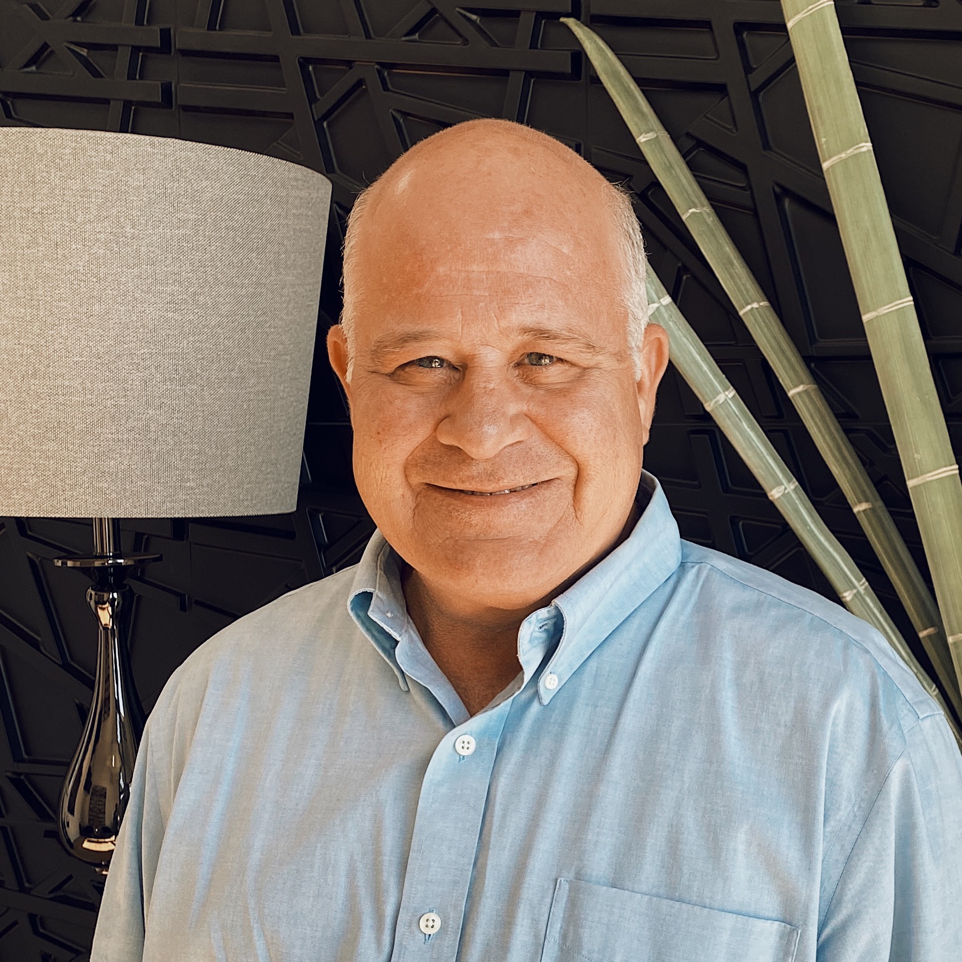 A smiling older man with a bald head and light blue button-up shirt stands indoors near a lamp and tall green plant against a black geometric-patterned wall.