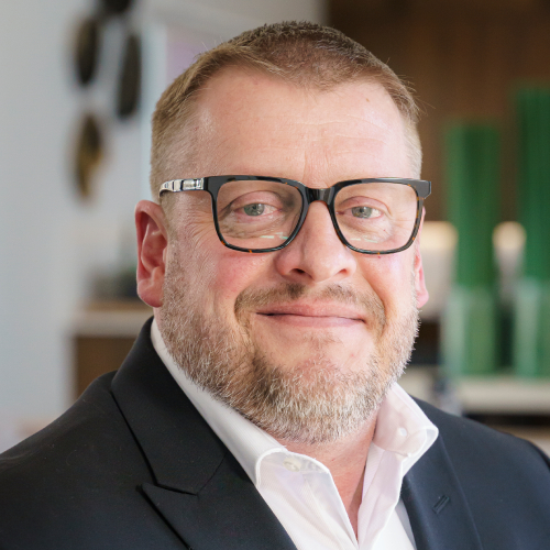 Brian-Dwiggins-2025-v2 A middle-aged man with light hair, a short beard, and glasses is smiling. He is wearing a dark suit jacket and a white shirt, with a blurred indoor background behind him.