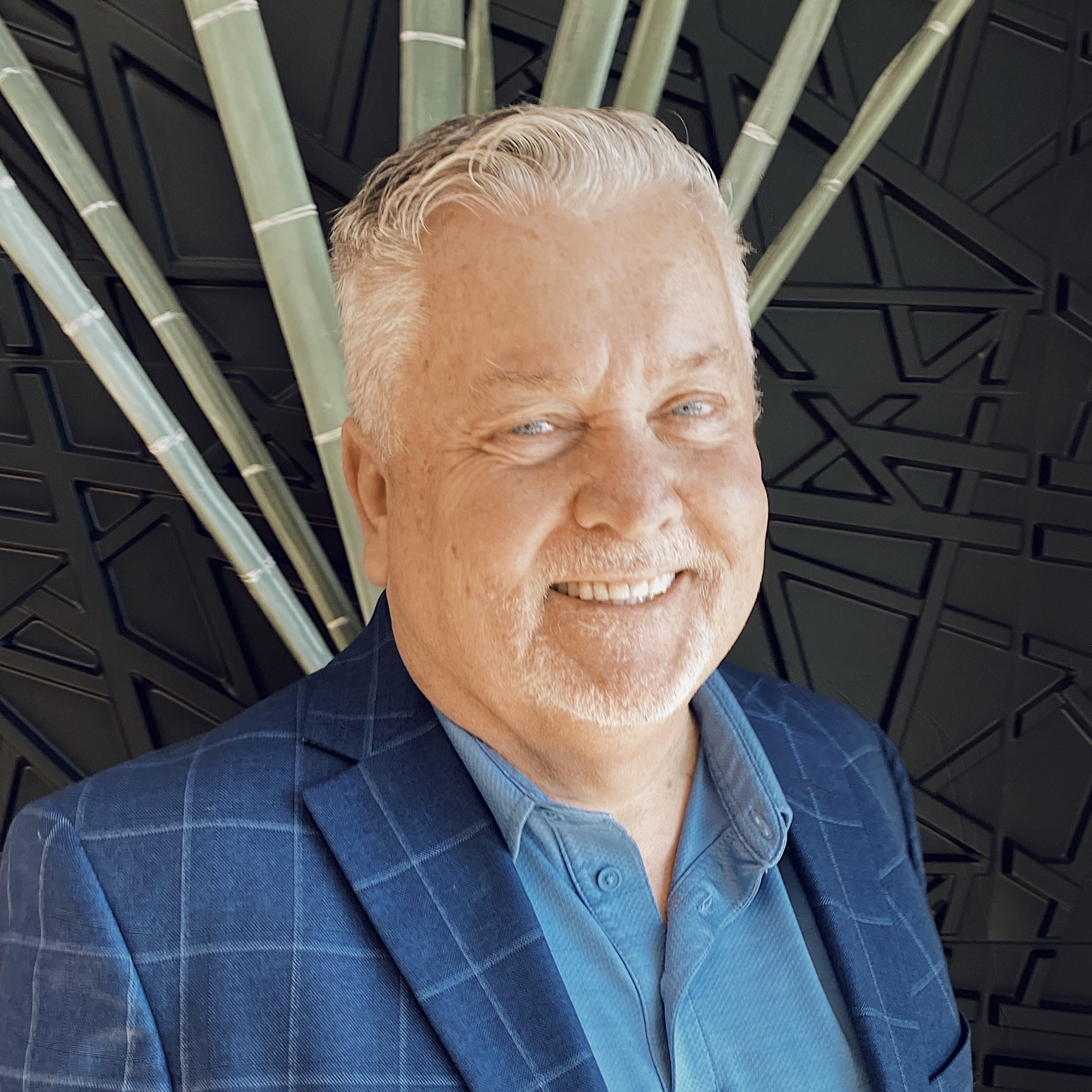 A smiling man with light hair and a short beard, wearing a blue plaid blazer and blue shirt, stands in front of a dark geometric wall with green bamboo-like plants behind him.
