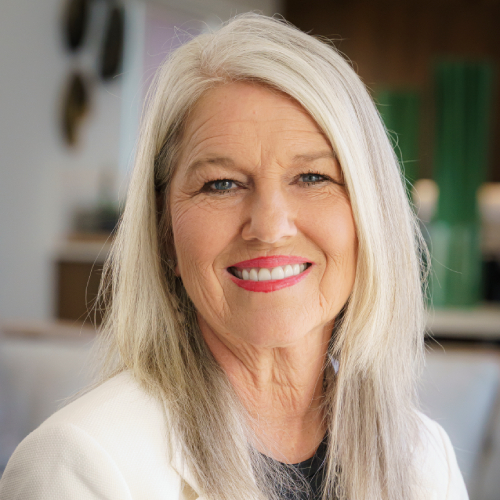 Susan-Long-2025-v2 Smiling older woman with long gray hair, wearing a white blazer and dark top, poses indoors with a blurred background featuring green decor and soft lighting.