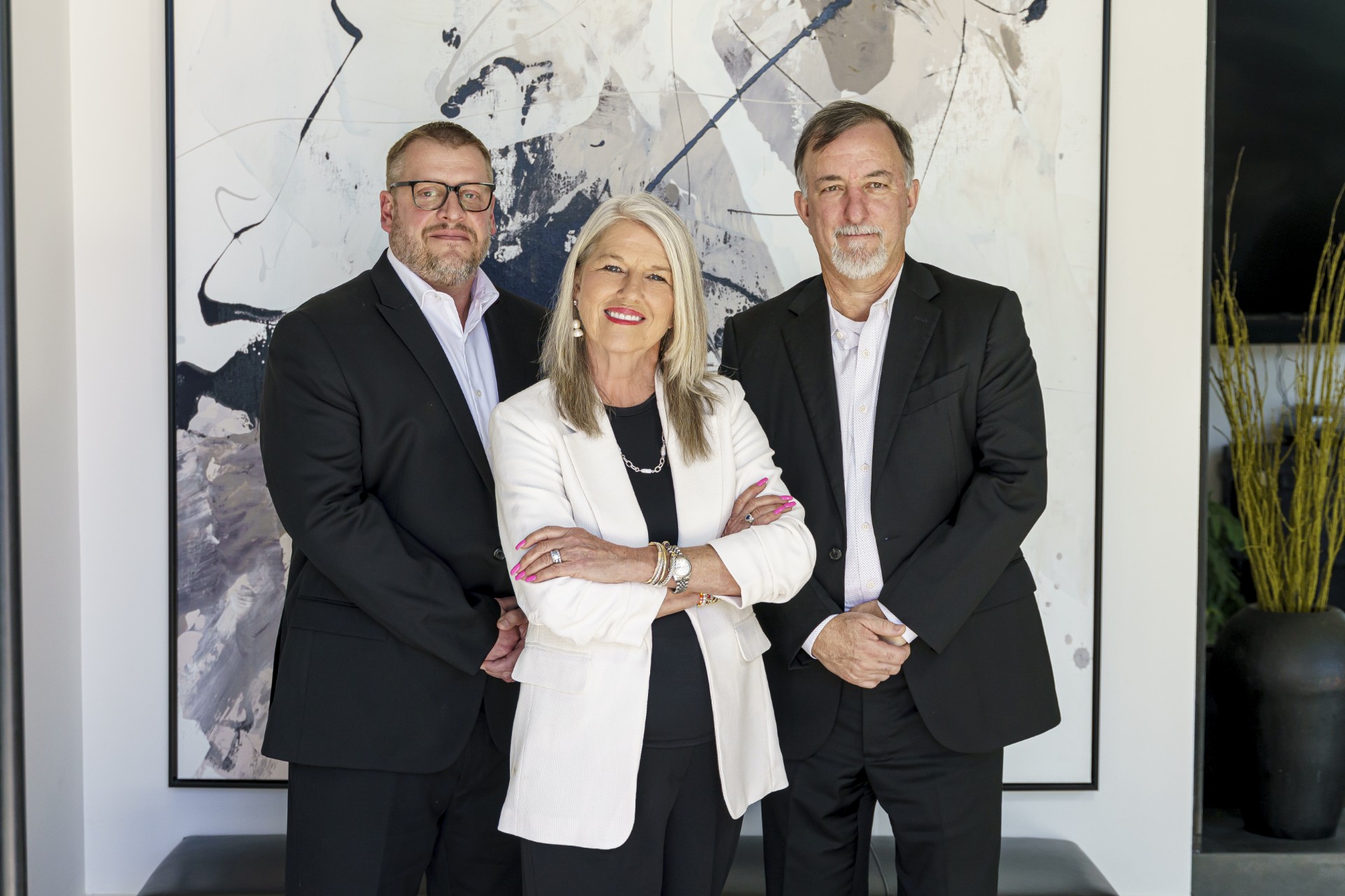 Trio Three professionally dressed adults stand together in front of an abstract black and white painting. The woman in the center wears a white blazer, flanked by two men in dark suits. All are smiling and facing the camera.