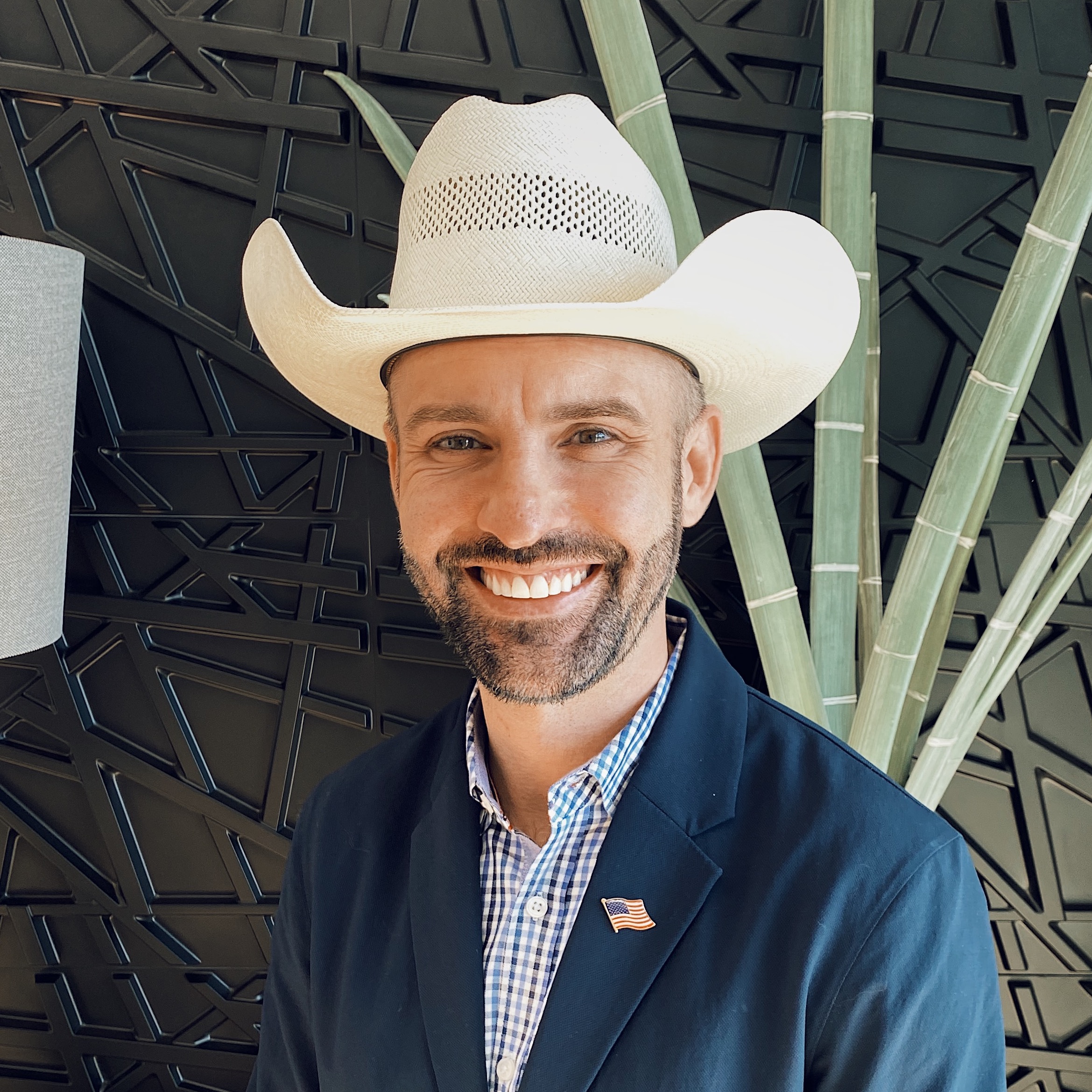 A smiling man in a white cowboy hat and blazer stands in front of a geometric black wall with green plant leaves. He wears a checkered shirt and an American flag pin on his jacket.