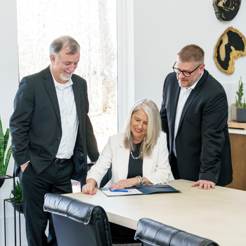 the-owners-updated Three business professionals in suits are gathered around a table in a modern office. A woman is seated, signing a document, while two men stand beside her, smiling and looking at the paperwork.