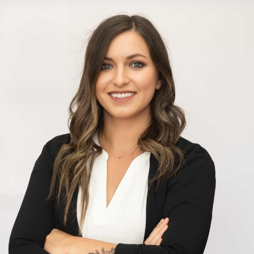 A woman with long wavy brown hair, wearing a white blouse and black blazer, stands smiling with her arms crossed against a plain light background.