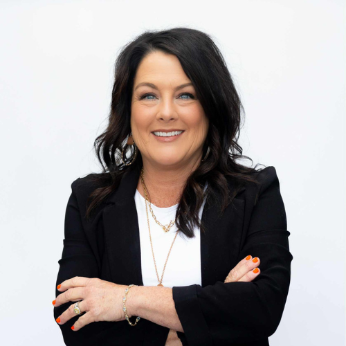 A woman with long dark hair, wearing a black blazer over a white top, stands with arms crossed and smiles at the camera. She has orange nail polish and gold jewelry. The background is plain white.