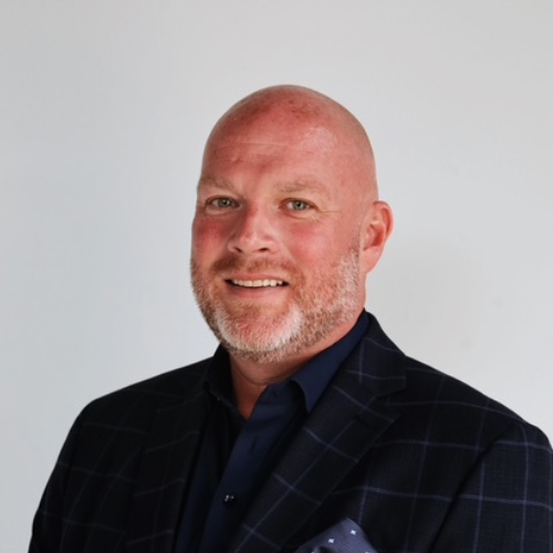 A bald man with a trimmed beard is smiling and wearing a dark checkered blazer over a navy shirt, standing against a plain light background.