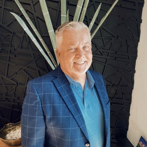 A smiling older man with white hair and a beard wears a blue checkered blazer over a light blue shirt, standing in front of a black textured wall with tall, green decorative leaves behind him.