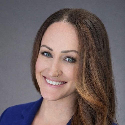 A woman with long brown hair, blue eyes, and a nose ring smiles at the camera. She is wearing a blue blazer and is posed against a plain gray background.