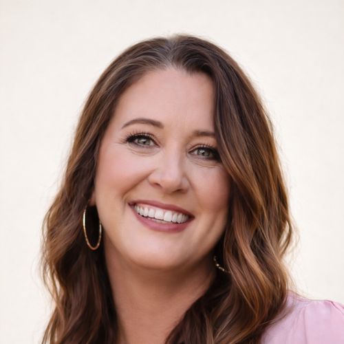 A woman with long, wavy brown hair and hoop earrings smiles at the camera. She is wearing light makeup, a pink top, and is posed in front of a plain, light background.