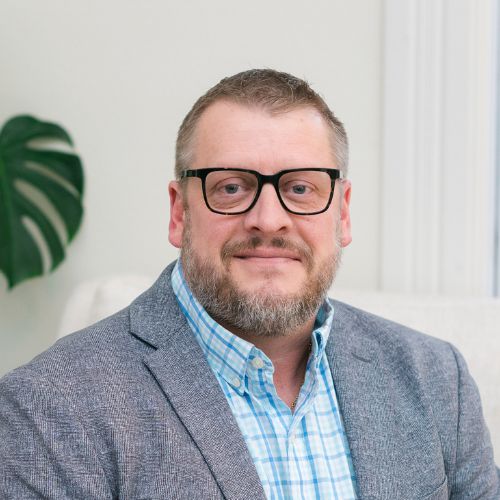 A man with short hair, a beard, and glasses wears a gray blazer over a blue checked shirt. He is seated indoors with a neutral background and a green plant visible.