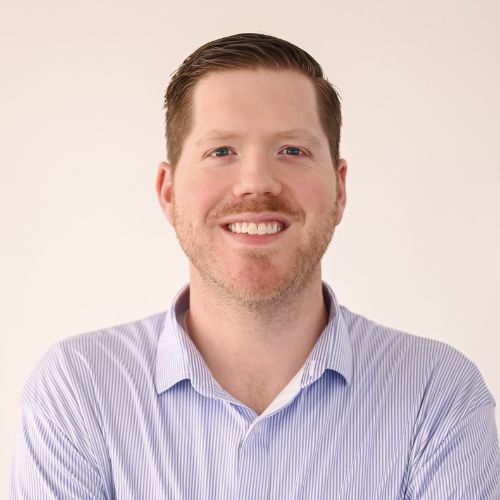 A man with short brown hair and a beard, wearing a light blue striped button-up shirt, smiles at the camera against a plain light background.