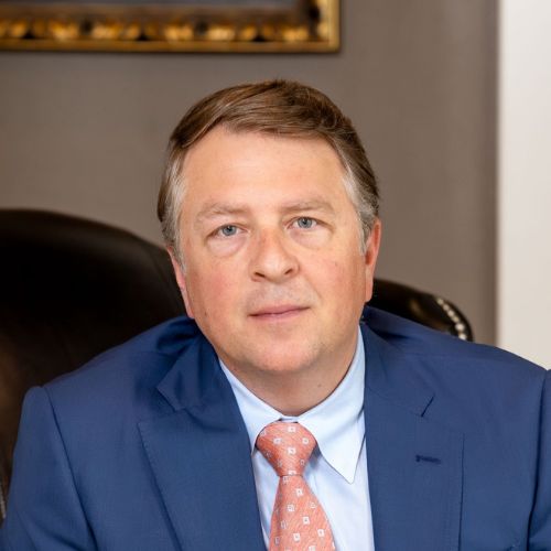 A middle-aged man with short, light brown hair is wearing a blue suit, light blue shirt, and peach patterned tie, seated in a dark leather chair, facing the camera in an indoor setting.