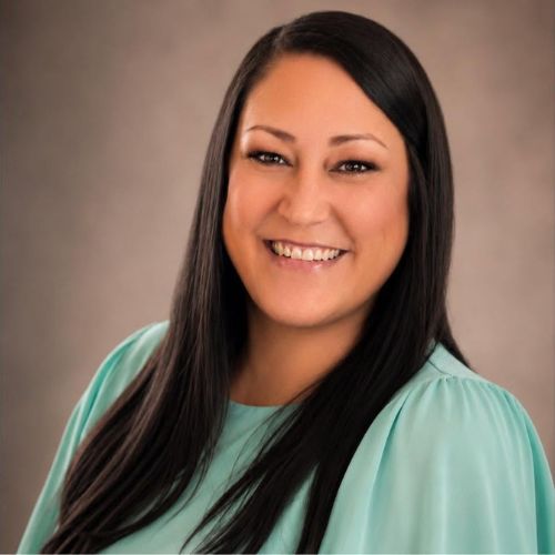 Smiling woman with long, straight dark hair wearing a light green blouse, posed in front of a plain brown background.